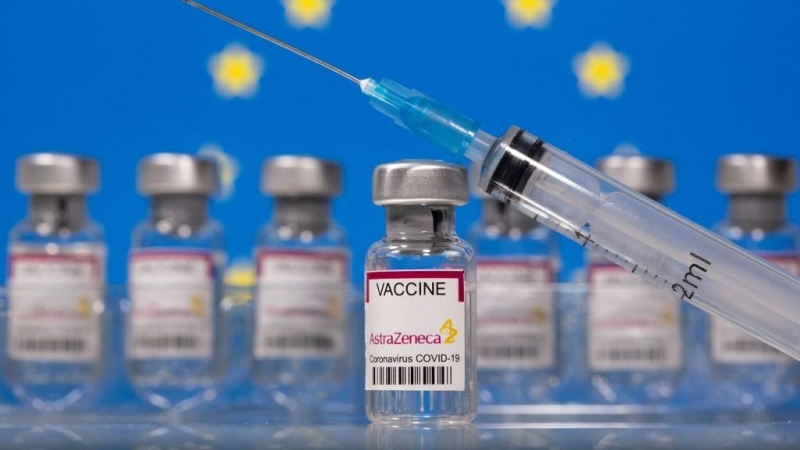 Un vial de vacunas etiquetado con el nombre de Astra-Zeneca, con una jeringuilla, y la bandera de la UE al fondo.