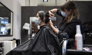 27/04/2020.- Una peluquera corta el pelo a una mujer con una máscara facial protectora contra el coronavirus en una peluquería de Berna, Suiza este lunes. Servicios como la peluquería, los masajes, los tatuajes o los salones de belleza están abiertos al p